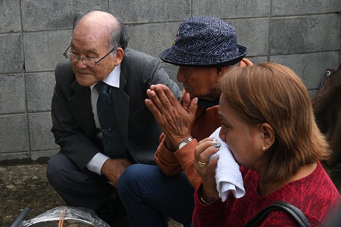 親族と思われる香村安紀さん（左）に説明を受け、香村家の墓に向かって手を合わせるアカヒジ・サムエルさん（中央）とオルミド・ミチコさん＝16日、うるま市の平安座葬祭場
